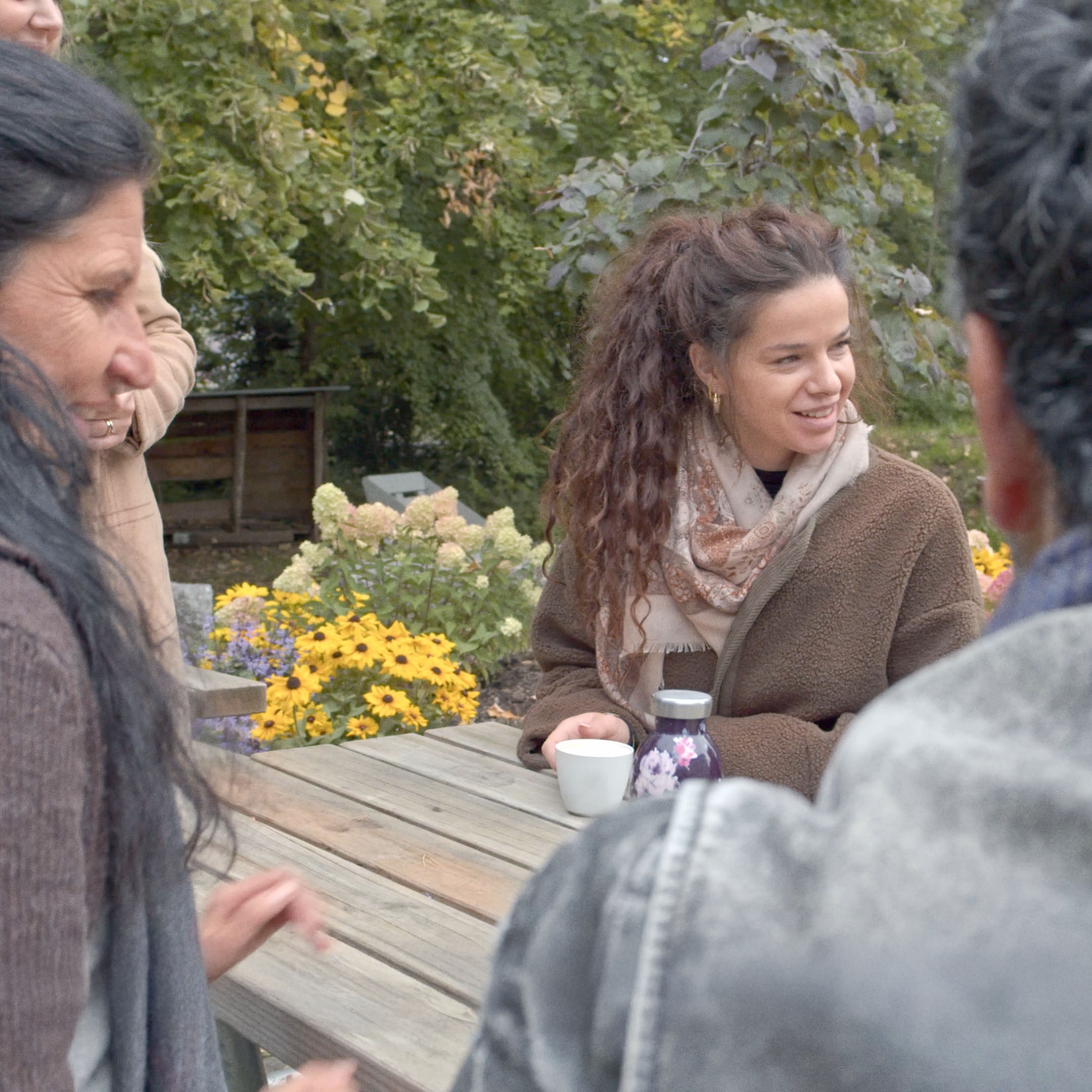 Group of people sitting around a wooden picnic table outdoors with yellow and purple flowers and greenery in the background.