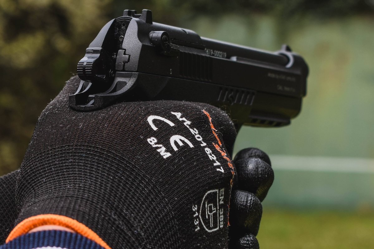 Close-up of a gloved hand holding a pistol against a blurred outdoor background.