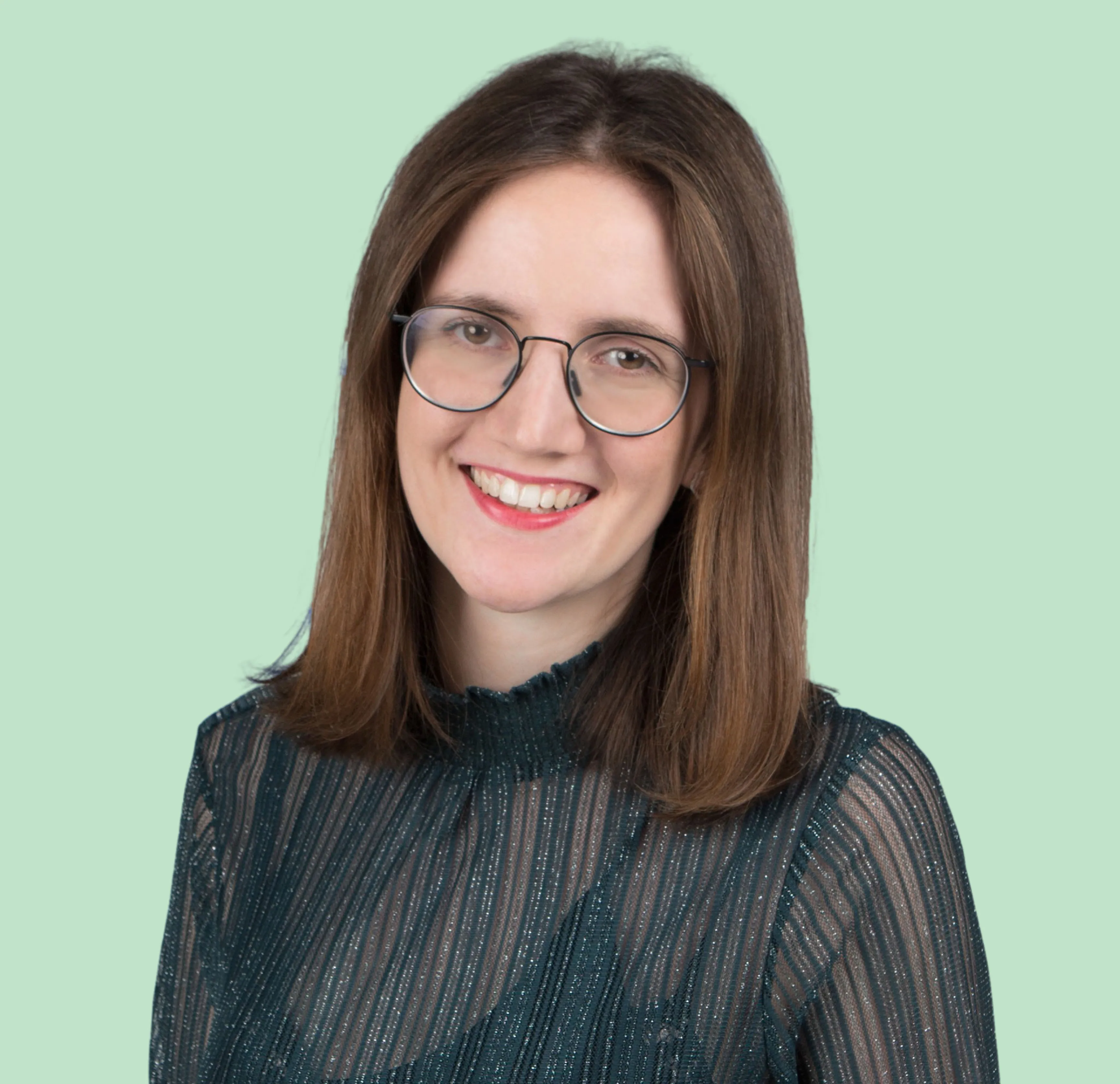 Rebecca Kremer schaut direkt in die Kamera und lächelt dabei Zähne zeigend. Diese Person hat schulterlanges Haar und trägt ein durchschimmerndes, gestreiftes schwarzes Oberteil, unter dem sie ein schwarzes Top darunter. Sie trägt eine Brille.