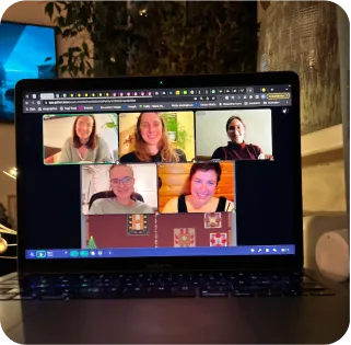 Eine Fotografie, auf der ein Laptop auf einem Tisch zu sehen ist. Auf dem Laptop Bildschirm sieht man, dass gerade ein Onlinemeeting zwischen fünf Personen stattfindet.
