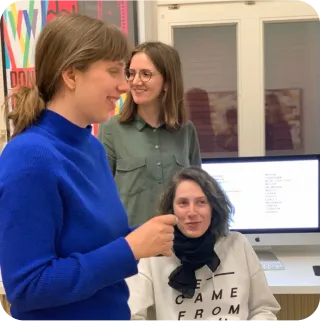 Ein Foto, auf dem drei Personen zu sehen sind. Die vordere ist nach rechts gedreht, hat einen Zopf und einen kräftigblauen Pullover. Unten sitzend ist eine weitere Person mit einem weißen Pullover. Dazwischen befindet sich die dritte, die eine Brille trägt und ein grünes Hemd. Sie befinden sich alle in einem Raum. Auf einem Tisch steht ein Monitor.