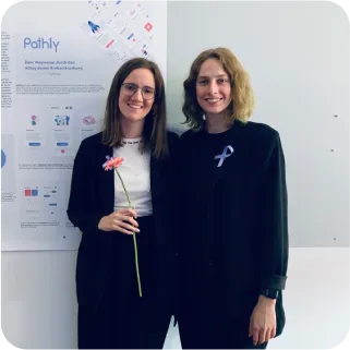 Links Rebecca und rechts Thora stehen vor einer Wand. Hinter den beiden sind große ausgedruckte Layout-Entwürfe von Pathly. Rebecca hält eine Blume in der Hand und Thora hat eine fliederfarbene Cancer-Ribbon auf der Brust.