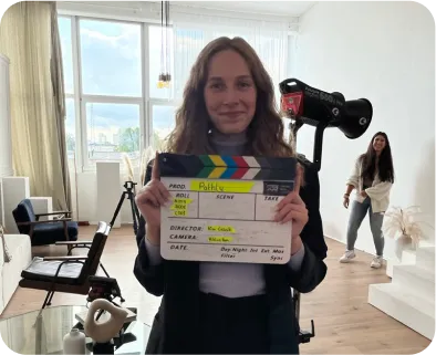 Eine Person steht in einem Raum in Richtung Kamera und lächelt dabei fröhlich. Diese hält außerdem eine Filmklappe in die Kamera. Im Hintergrund ist eine weitere Person zu sehen, die auch in die Kamera lächelt.