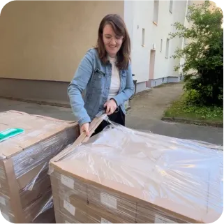Eine Person namens Rebecca steht vor einer großen Lieferung grauer Kartons, die sie glücklich lächelnd öffnet. Dabei steht sie draußen. 