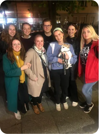 Acht Personen stehen draußen in der Nacht einer Großstadt. Alle lächeln in die Kamera. Die dritte Person von links, die in der vorderen Reihe steht, hat einen weißen Hund mit braunen Flecken auf dem Arm.