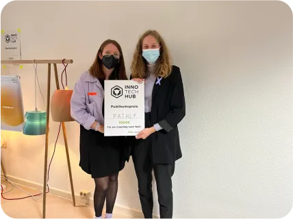 Eine Fotografie zweier Personen, die nebeneinanderstehen und zwischen sich eine große Preistafel halten. Auf dieser ist folgendes zu lesen: INNO TECH HUB Publikumspreis Pathly 1000€. Beide Personen tragen medizinische Masken.
