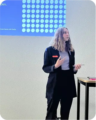 Foto einer Person, die gerade dabei ist, einen Vortrag zu halten. Hinter dieser Person befindet sich eine weiße Wand, außerdem kann man oben links im Bild erkennen, dass dort die Präsentationsfolien zu sehen sind. Vor dieser Person steht ein Tisch. Die Person trägt ein schwarzes Sacco mit einem weißen Oberteil darunter und eine schwarze Hose. Ihr Haar ist schulterlang, wellig.