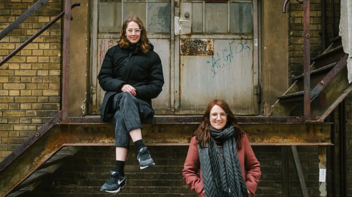 Fotografie zweier Personen. Beide sind im Vordergrund eines Fabrikgebäudes mit Stahltreppe. Die Linke Person sitzt auf der Stahltreppe und die rechte steht. Beide Personen tragen warme Jacken.