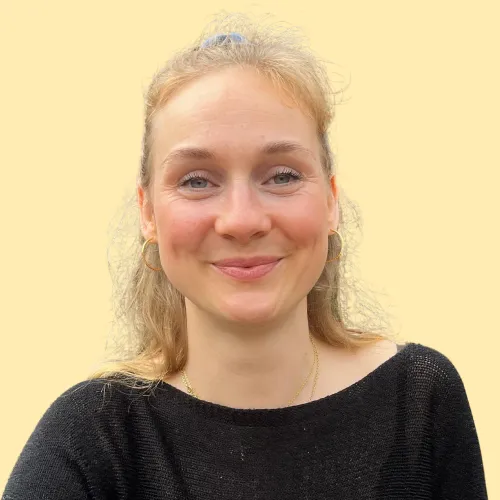 Marie hat Blonde lockige Haare und ein schwarzes Oberteil. Sie steht vor einem gelben Hintergrund.