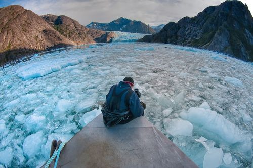 A photographer sitting on the front of a small boat taking video of a seascape filled with floating ice all around him.