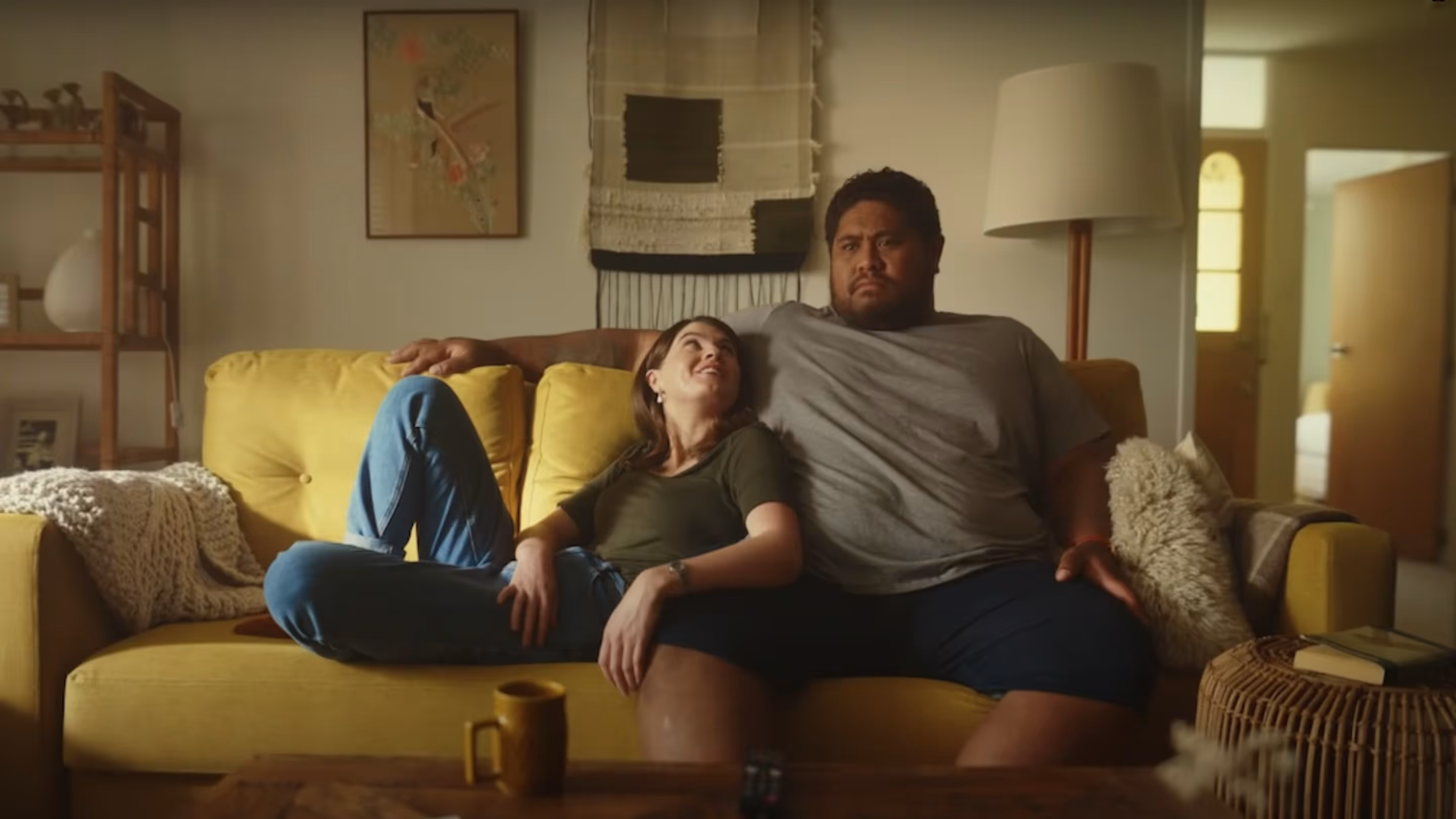 A couple are cuddling on a yellow couch and looking at each other in their cluttered living room.
