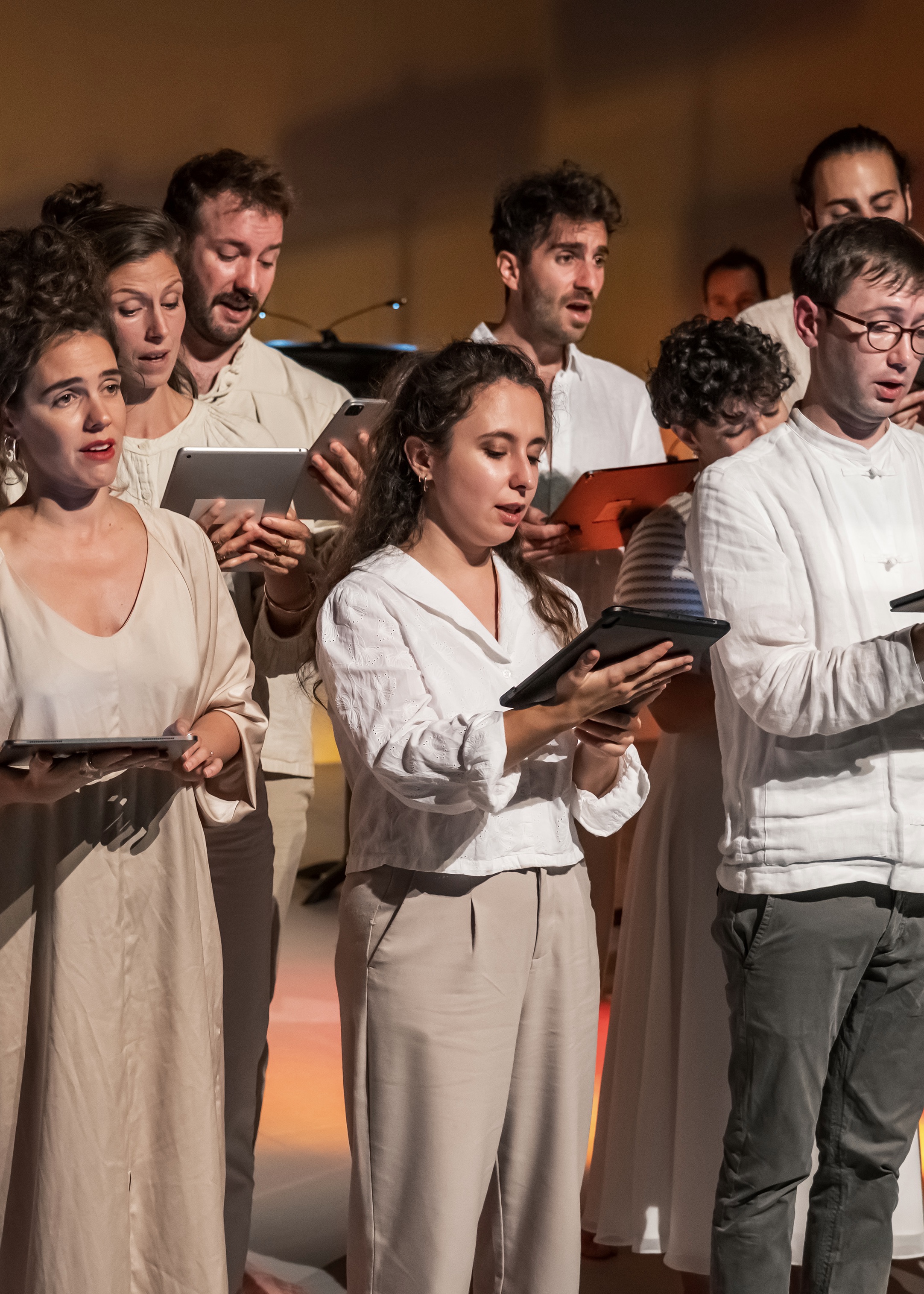 Ein Vokalensemble und Dirigent im Vordergrund, alle in hellen Farbtönen gekleidet und in warmes Licht getaucht