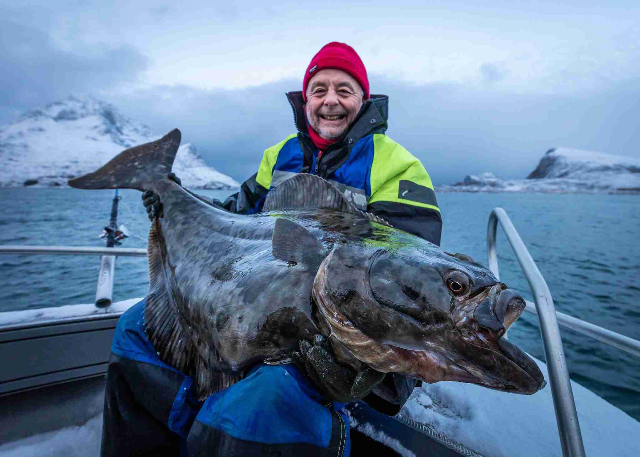 Smilende fisker med stor kveite om bord i båt utenfor Henningsvær, omgitt av snødekte fjell og kaldt vinterhav.