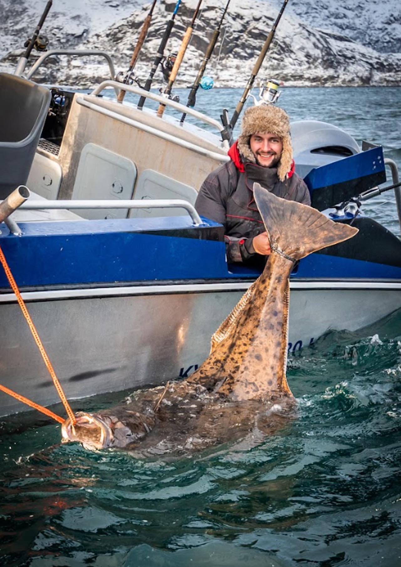 Man holding a large halibut from a winter fishing trip in Lofoten, next to a boat in Arctic waters.