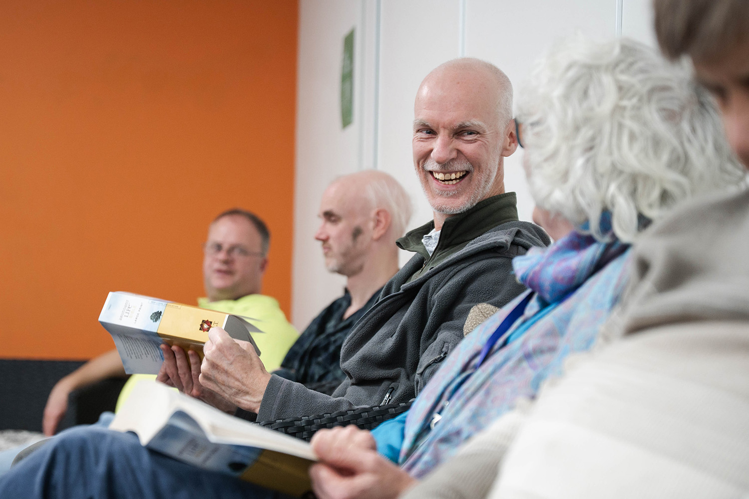 A group of adults sitting in a row indoors, reading books and smiling, with an orange wall in the background.
