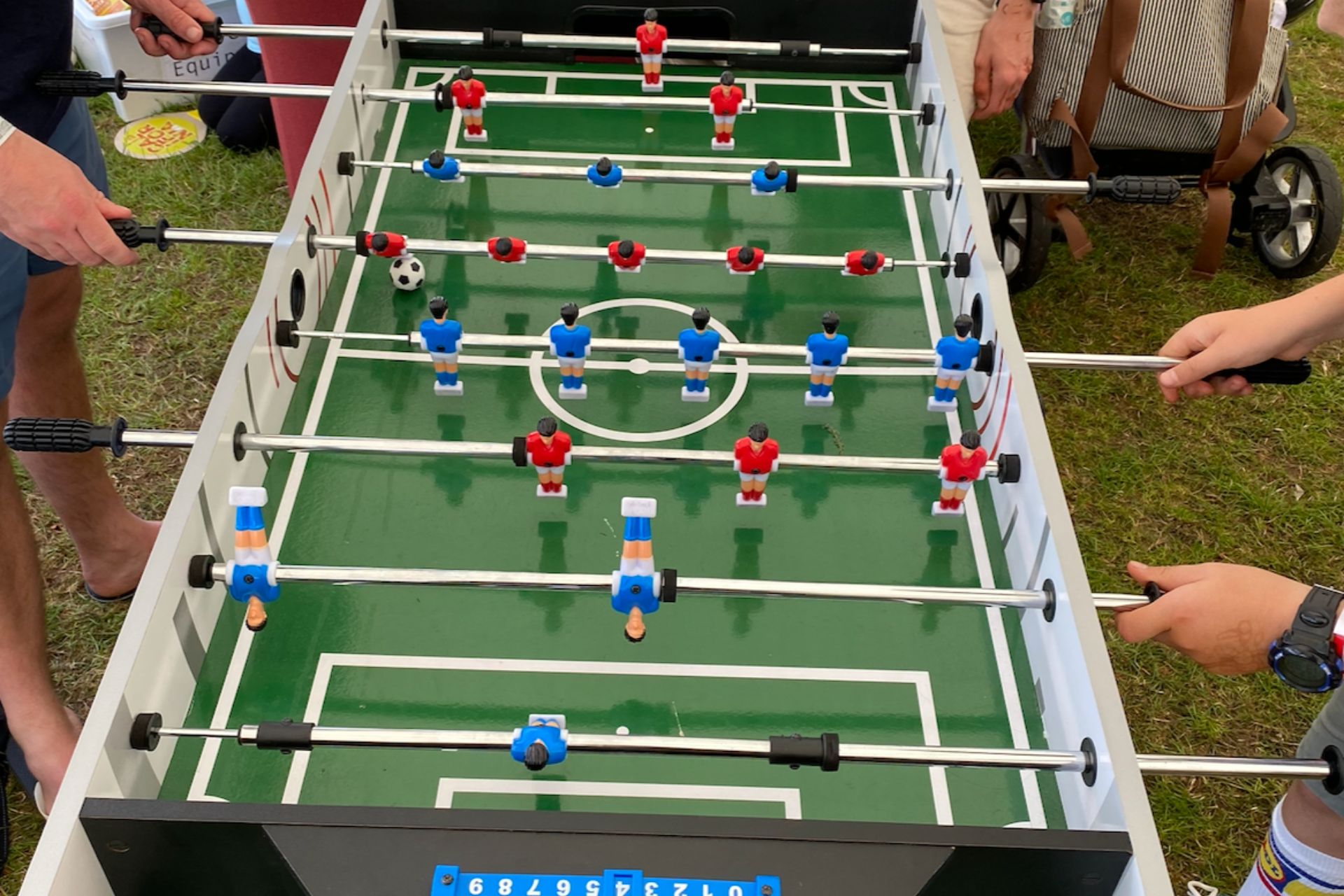 Two people playing foosball outdoors on a green foosball table with red and blue players, with a foosball ball near the red player's row.