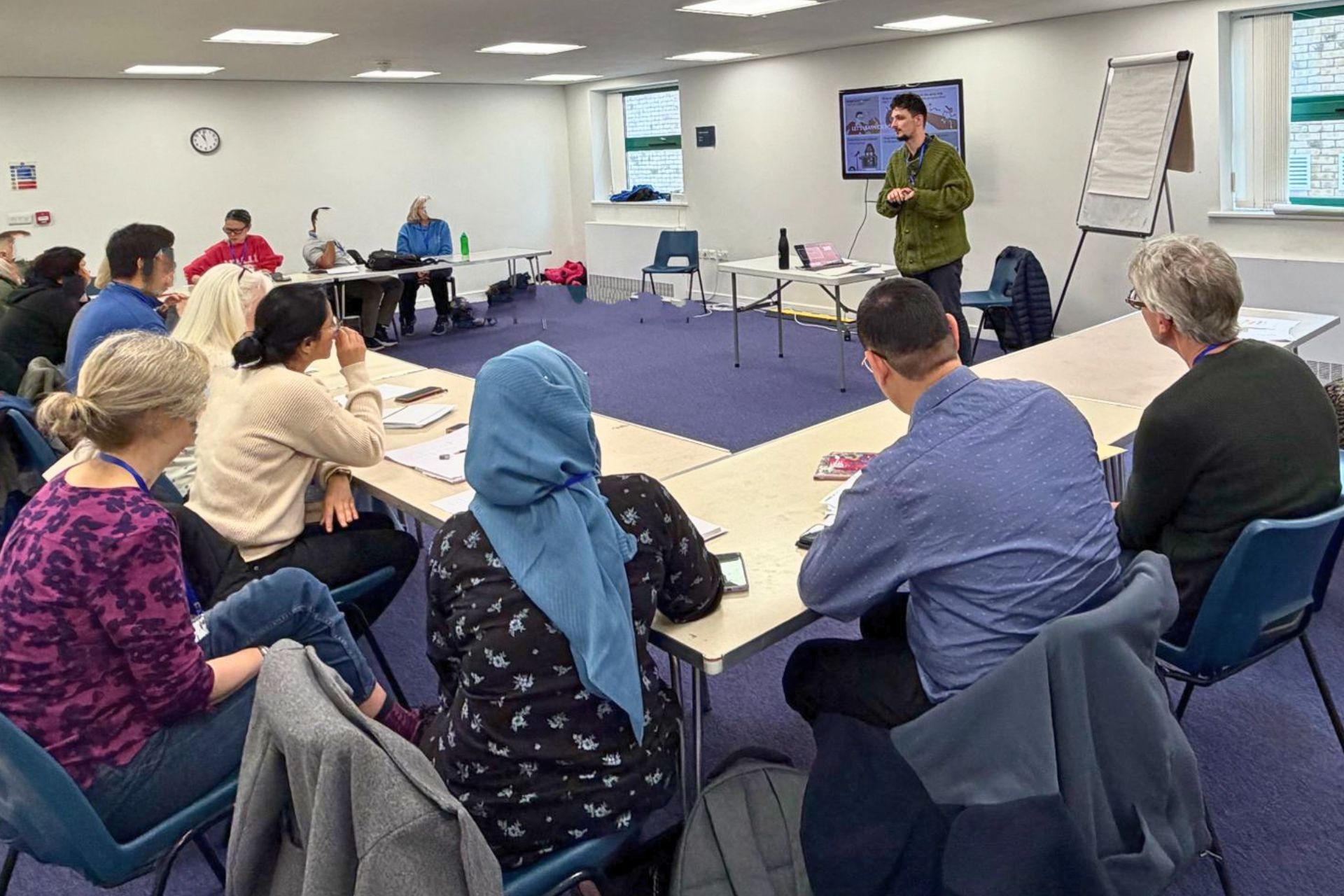 Five diverse young adults sitting around a table having a friendly discussion with coffee mugs in an informal indoor setting.