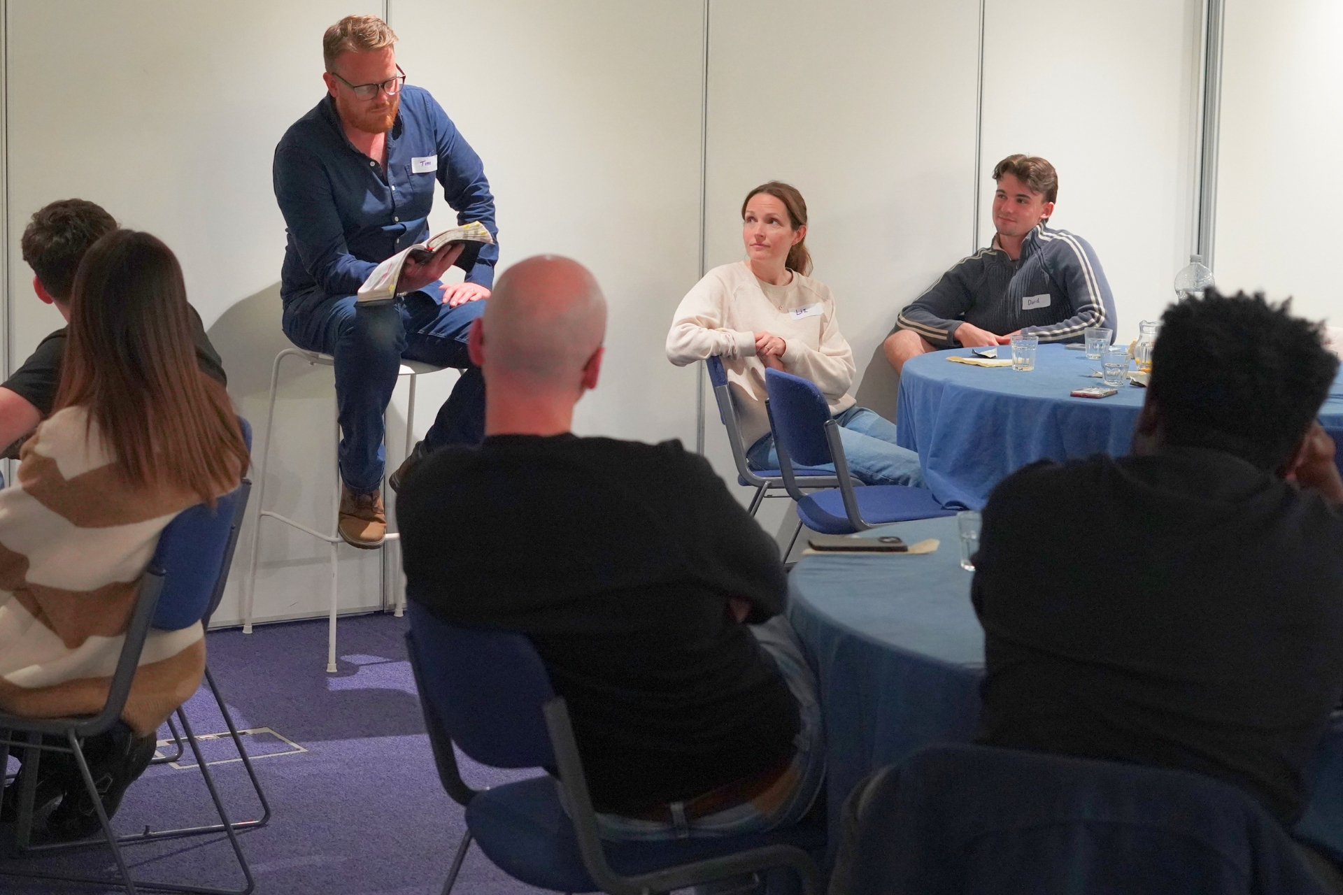 Man with glasses reading from a book to a seated group in a casual meeting room.