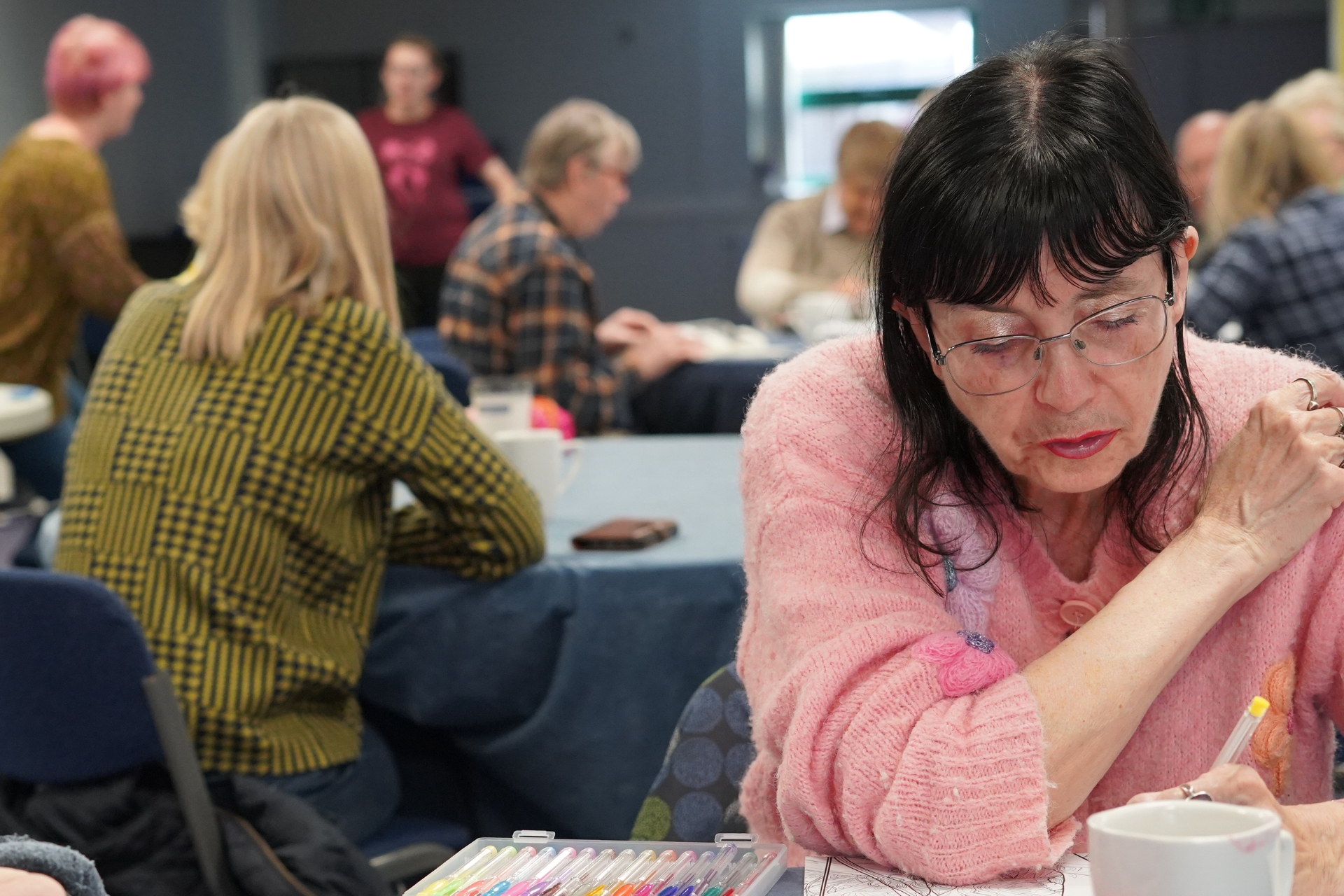 A woman in a pink sweater and glasses focused on coloring with markers at a table in a group setting.