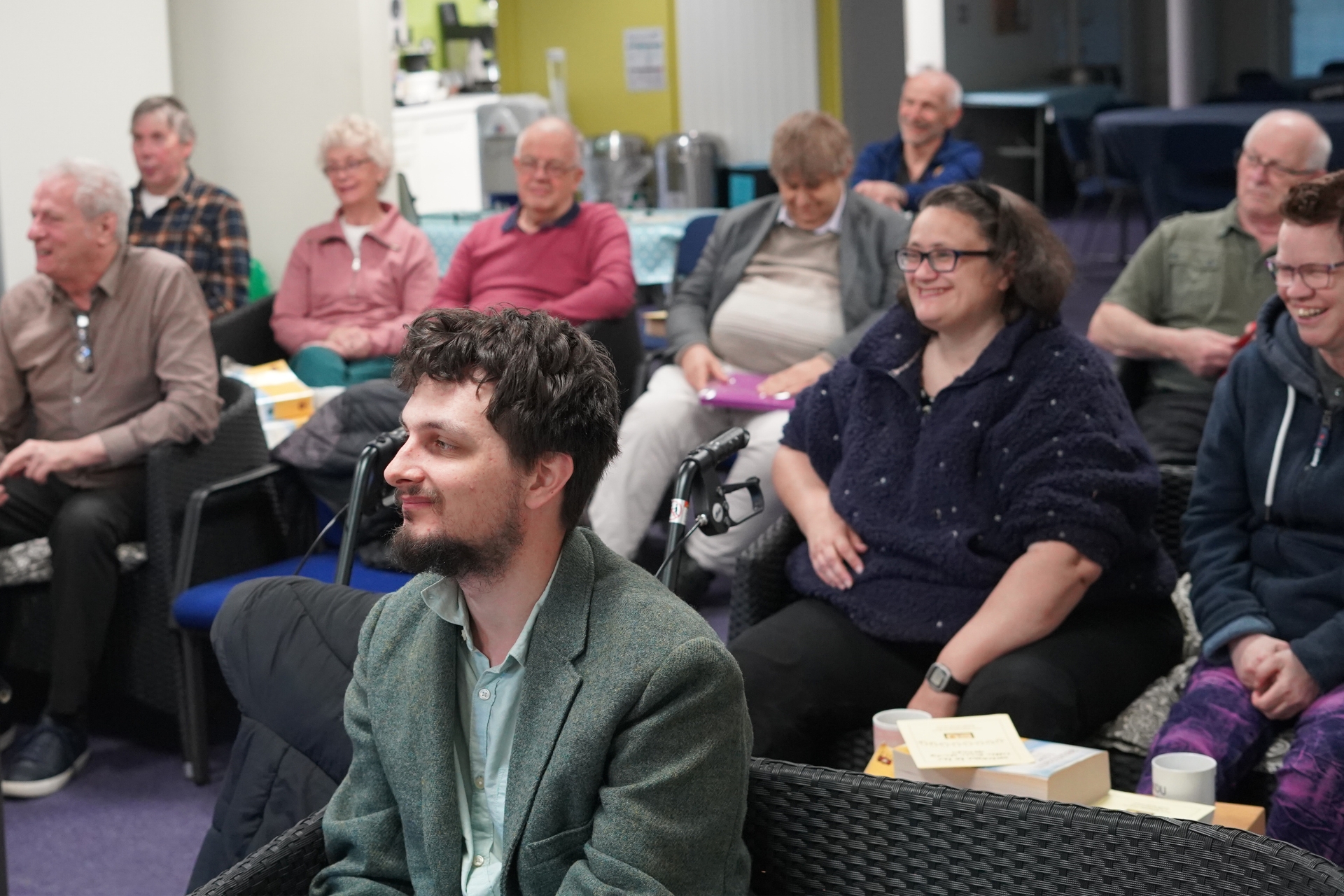 A group of people seated indoors, attentively listening with smiles and relaxed postures.