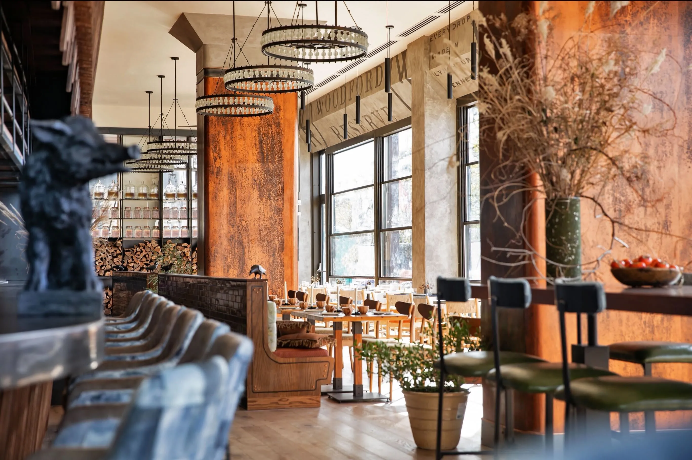 Bright modern restaurant interior with large windows, wooden tables and chairs, green bar stools, and rustic orange walls.
