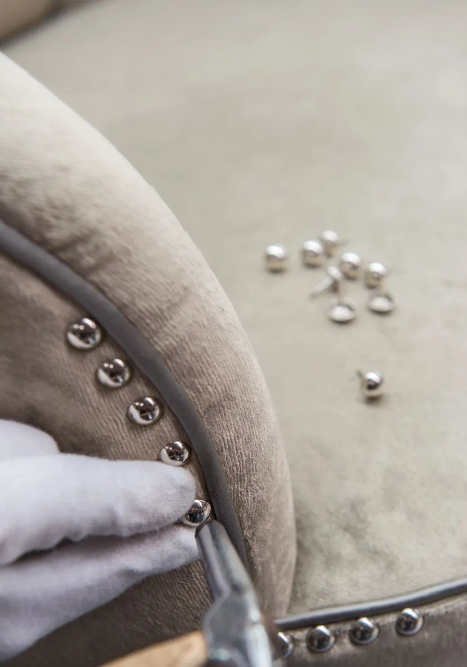 Close-up of a gloved hand using a hammer to attach decorative silver upholstery tacks to beige fabric furniture.