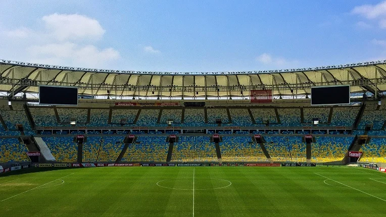 Vista panorámica desde el campo de juego de un imponente estadio de fútbol vacío, con gradas pintadas de azul y amarillo bajo una gran estructura de techo moderna. La imagen ilustra la magnitud de los escenarios deportivos listos para albergar los partidos del Mundial 2026.