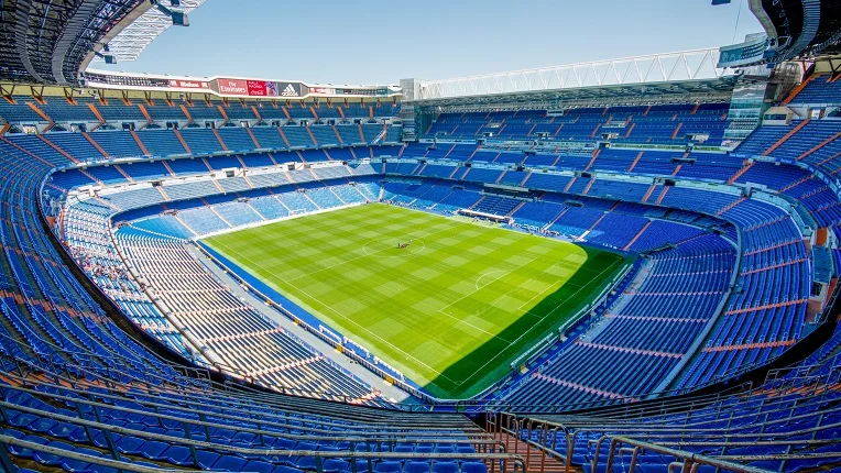 Vista aérea de un gran estadio de fútbol con un césped impecable y gradas vacías dispuestas en múltiples niveles de asientos de color azul. La imponente arquitectura deportiva representa los escenarios de vanguardia preparados para recibir la pasión del Mundial 2026.
