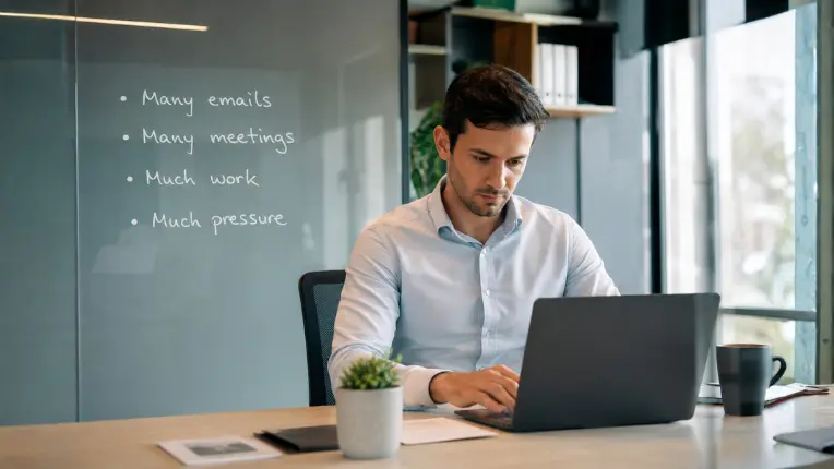 Profesional joven trabajando en una oficina moderna frente a un computador portátil, con un tablero de vidrio donde se leen ejemplos de sustantivos contables e incontables inglés: "Many emails", "Many meetings", "Much work" y "Much pressure".
