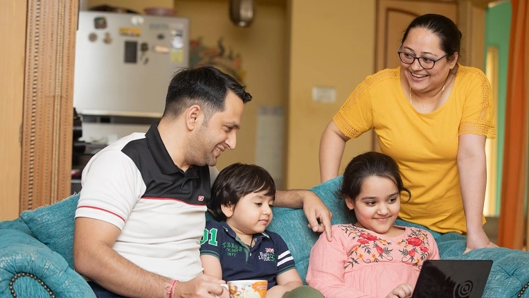 Una familia de cuatro miembros, está sentada en un sofá azul en su sala de estar. Todos miran sonrientes la pantalla de una tableta que sostiene la hija, sugiriendo la visualización de un curso de inglés online para niños, mientras el padre sostiene una taza.