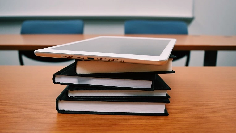 Una tableta digital colocada sobre una pila de libros de tapa negra en un escritorio de madera dentro de un salón de clases, representando las herramientas de estudio digitales y los cursos de inglés online para niños y adolescentes.