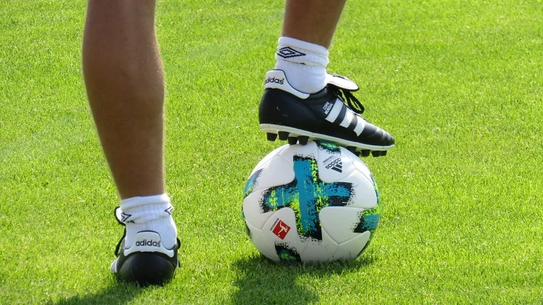 Primer plano de las piernas de un futbolista en un campo de césped verde, con un pie apoyado sobre un balón de fútbol Adidas blanco con detalles verdes y azules, ideal para ilustrar vocabulario de fútbol en inglés.