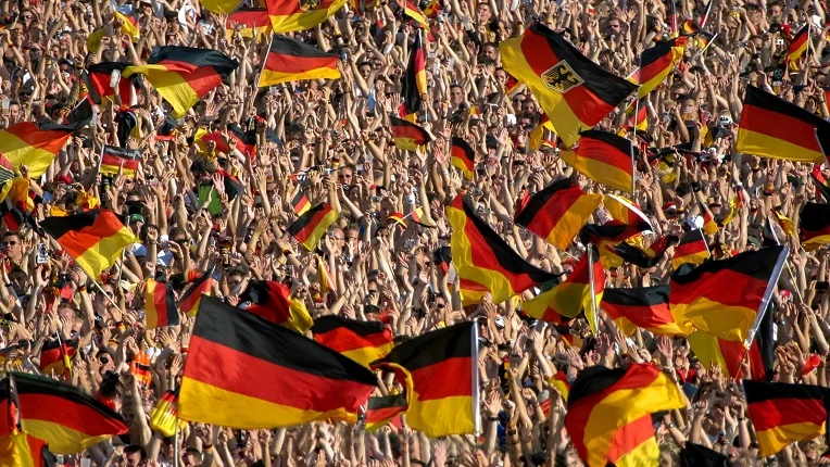 Una gran multitud de aficionados al fútbol en las gradas de un estadio, con los brazos en alto y ondeando numerosas banderas de Alemania, ideal para ilustrar el vocabulario de fútbol en inglés relacionado con la afición y el apoyo a un equipo.