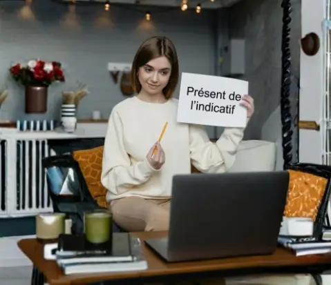 Una joven sostiene un cartel que dice "Présent de l'indicatif" frente a su laptop durante una clase en línea, representando el aprendizaje del presente simple en francés.