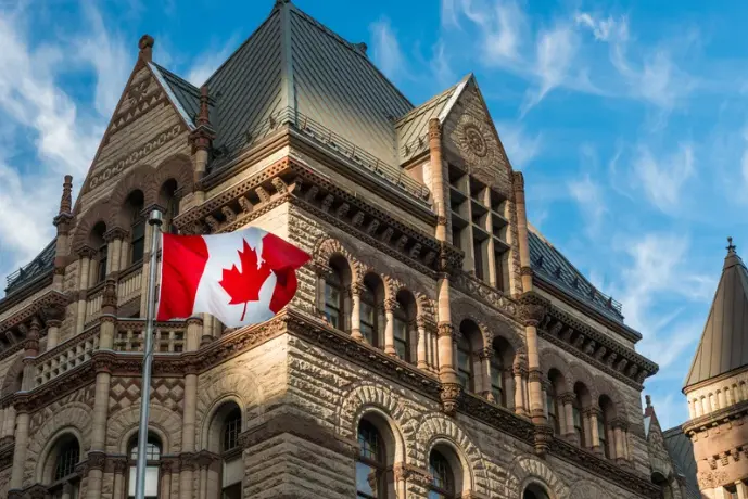La bandera de Canadá ondeando frente a la fachada de un edificio gubernamental histórico de piedra, ilustrando los procesos migratorios y el requisito de francés para migrar a Canadá.