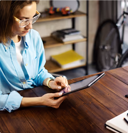 Woman wearing glasses and a blue shirt using a tablet at a wooden table with shelves and a bicycle in the background.