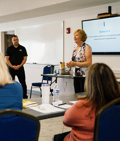 Woman standing and speaking in front of a classroom with seated attendees, a man observing beside a whiteboard and a presentation slide titled 'Scenario 4' displayed on a screen.