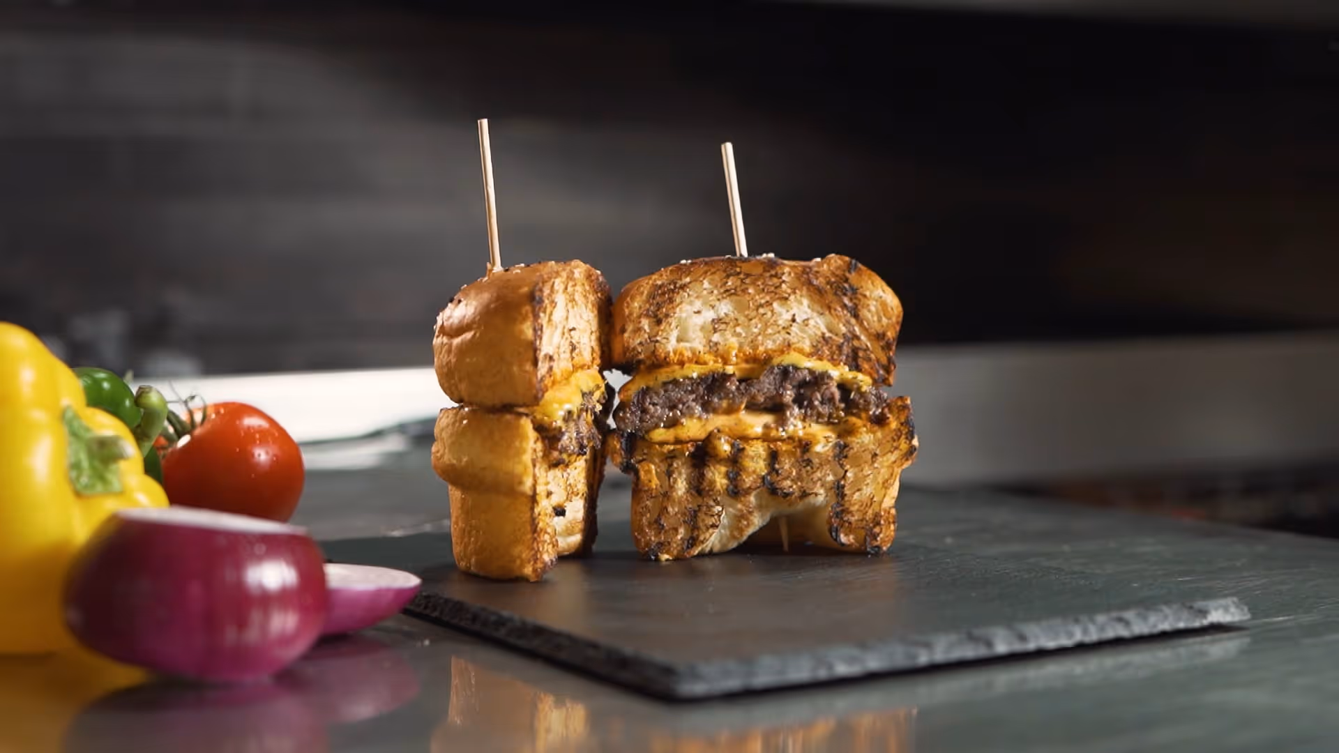 Gegrillte Cheeseburgerhälften mit geschmolzenem Käse auf schwarzer Schieferplatte, umgeben von frischem Gemüse.