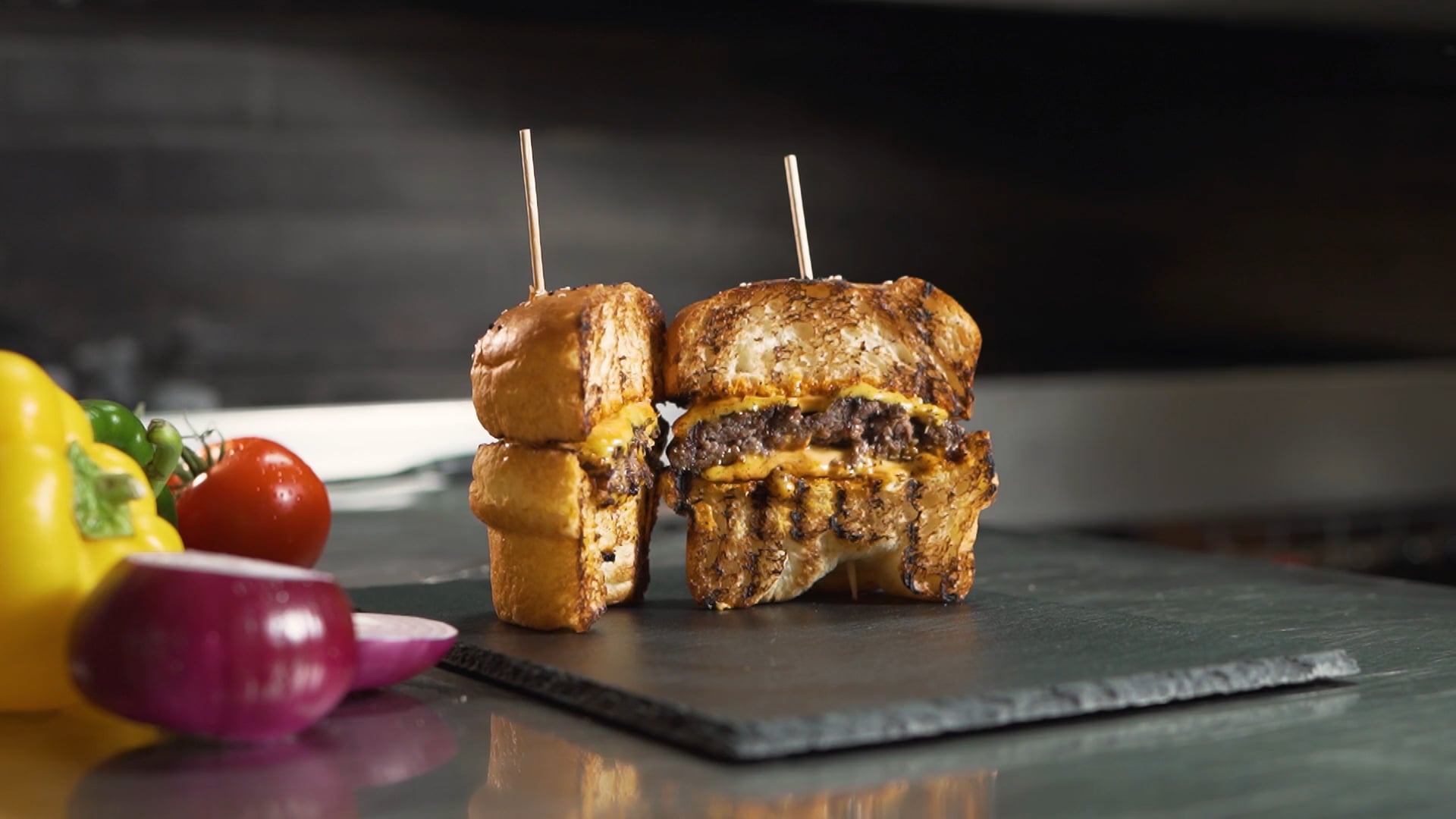 Gegrillte Cheeseburgerhälften mit geschmolzenem Käse auf schwarzer Schieferplatte, umgeben von frischem Gemüse.