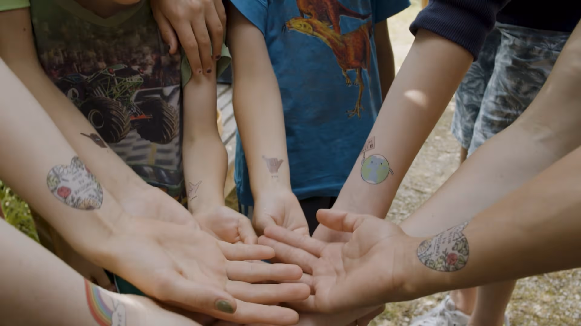 Mehrere ausgestreckte Arme von Kindern mit bunten temporären Tattoos, darunter Herzen, eine Weltkugel und eine Handgeste.