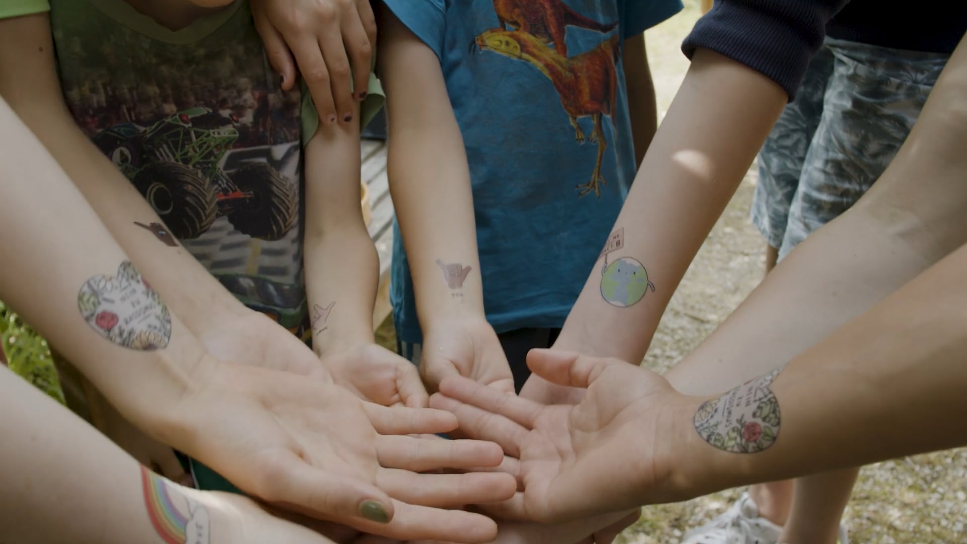Mehrere ausgestreckte Arme von Kindern mit bunten temporären Tattoos, darunter Herzen, eine Weltkugel und eine Handgeste.