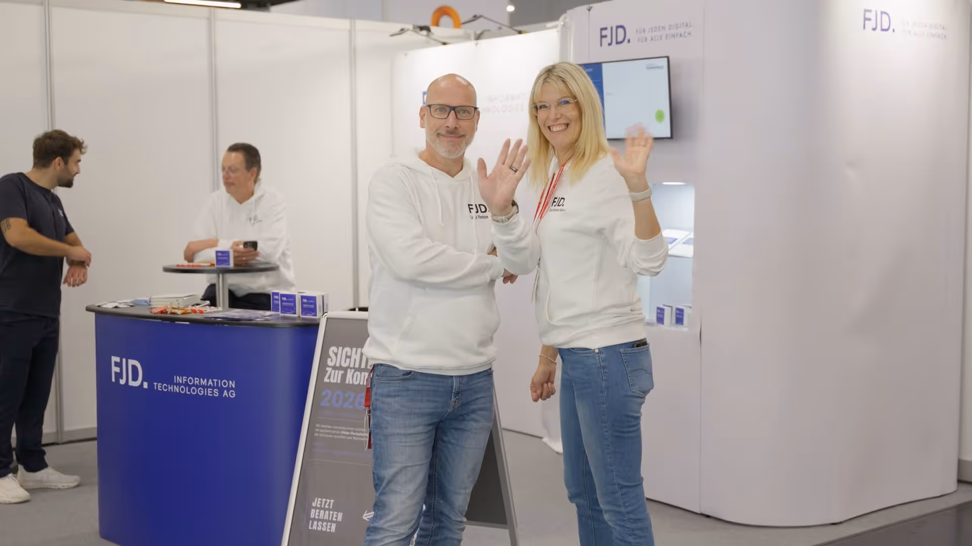 Zwei Personen in weißen FJD-Pullovern winken lächelnd in einer Messehalle mit blauem FJD-Informationstresen.