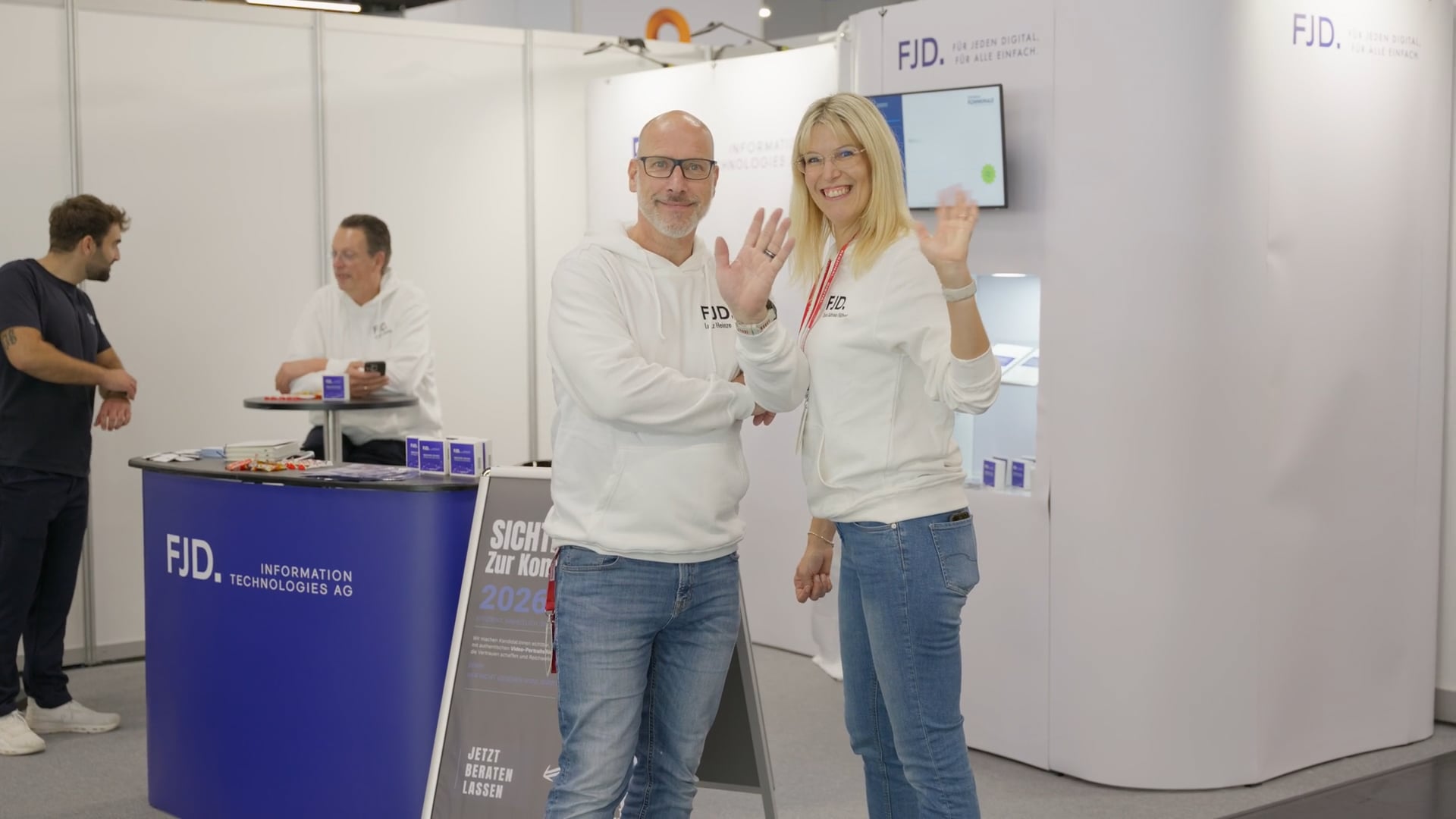Zwei Personen in weißen FJD-Pullovern winken lächelnd in einer Messehalle mit blauem FJD-Informationstresen.