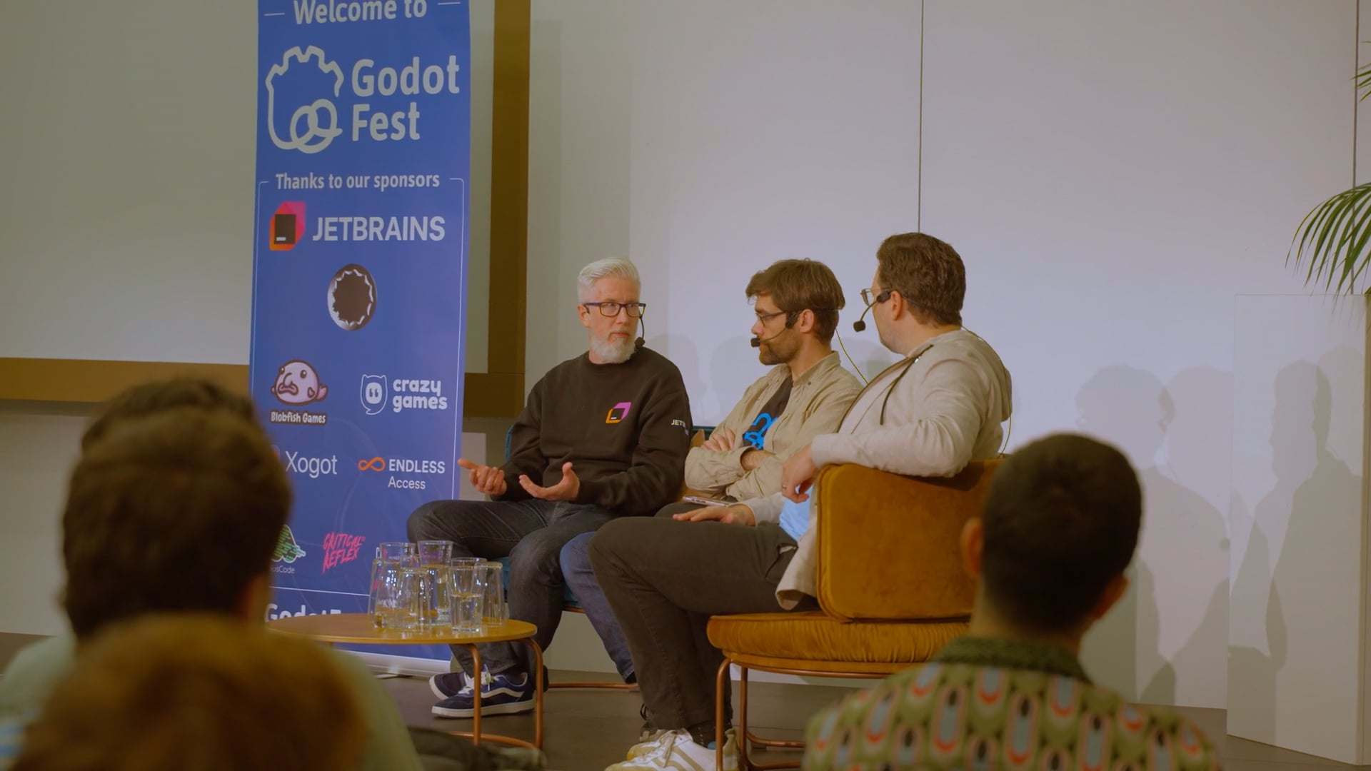 Drei Männer mit Headsets sitzen auf einer Bühne und führen bei der Godot Fest-Konferenz eine Podiumsdiskussion.