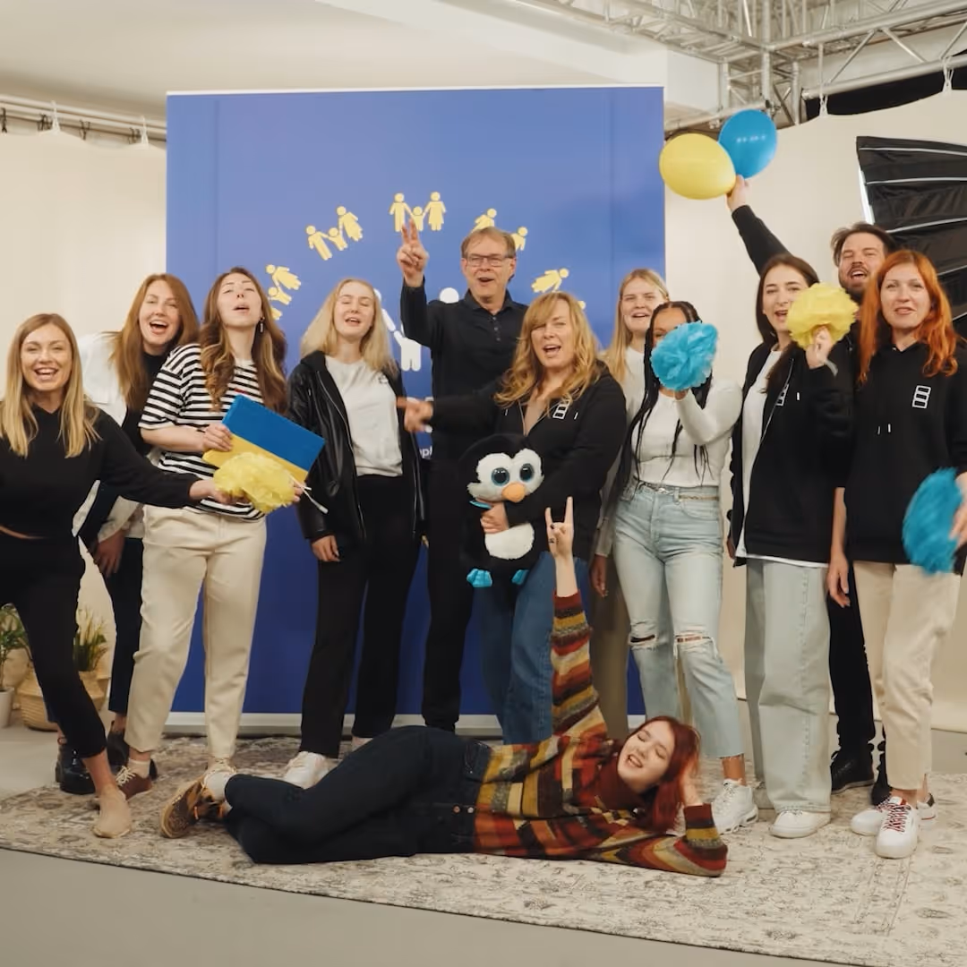 Gruppe von Menschen posiert lachend vor blauem Hintergrund mit gelben Figuren, einige halten blaue und gelbe Pompons, eine Person liegt auf dem Boden.