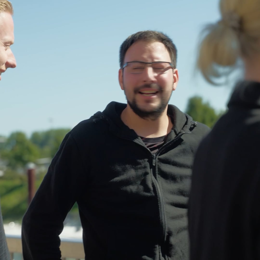 Drei Personen stehen im Freien im Gespräch, ein Mann in der Mitte lacht mit geschlossenen Augen.