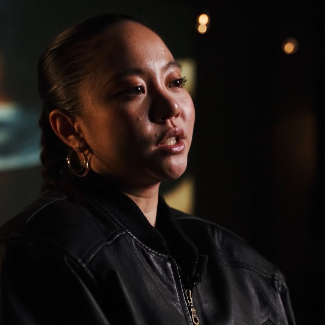 Frau mit zusammengebundenem Haar und großen Ohrringen, die eine schwarze Lederjacke trägt und im Halbdunkel spricht.