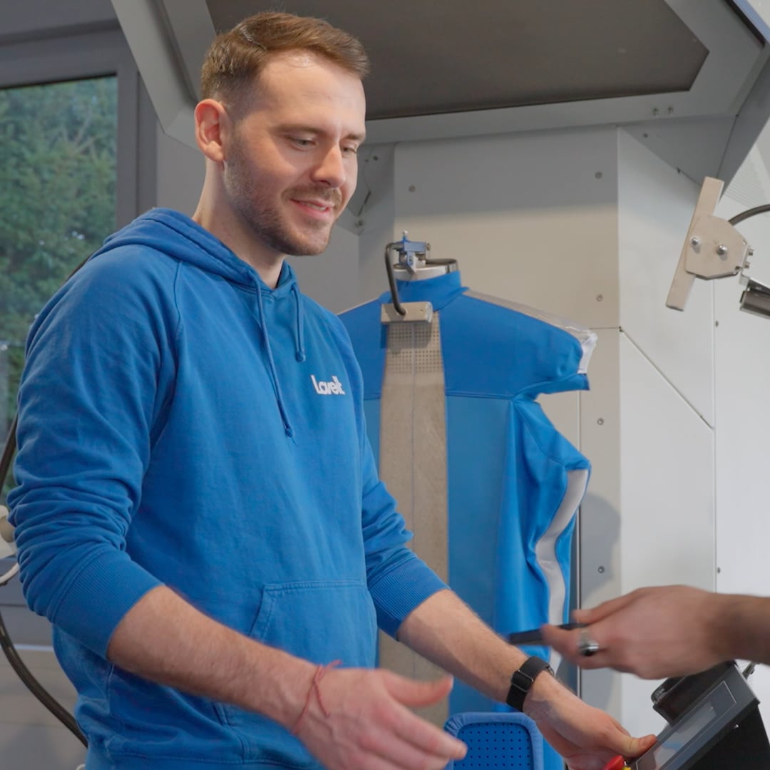 Ein lächelnder Mann in blauem Kapuzenpullover bedient eine Maschine in einem Raum mit technischem Gerät und einem Schaufensterpuppe mit blauem Überzug.