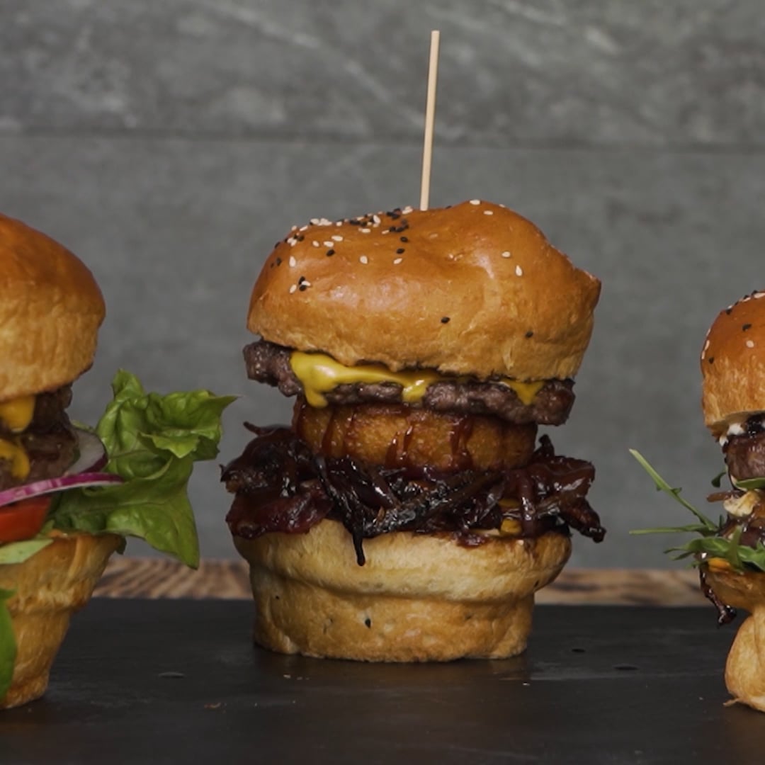 Burger mit Sesambrötchen, Rindfleischpatty, geschmolzenem Käse, Zwiebelringen und karamellisierten Zwiebeln auf dunklem Untergrund.