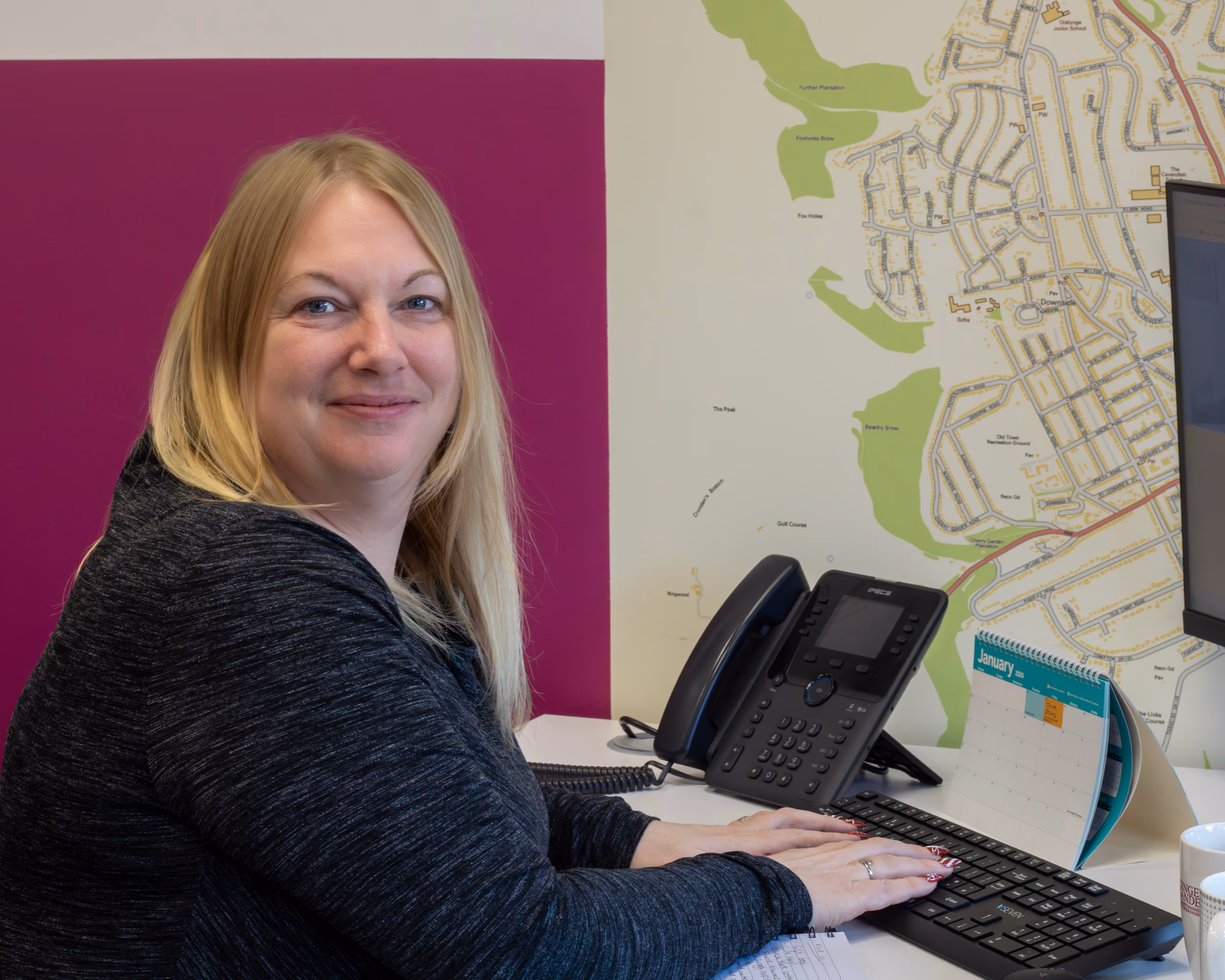 A woman sits at a desk using a computer, with a wall map visible behind her.